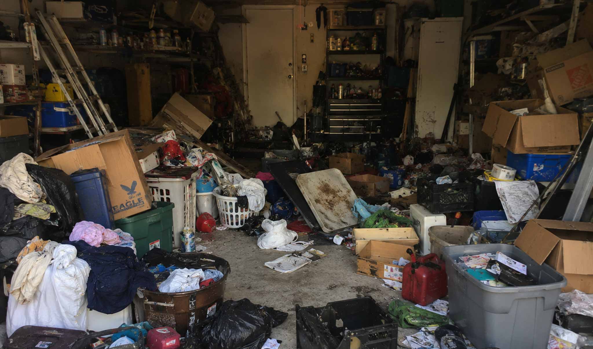 A messy and cluttered single-entry residential garage in St. George Utah packed with old boxes, plastic bins, and broken tools against a solid back wall.